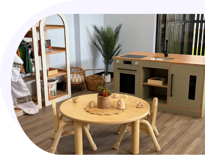 A modern childcare center play area featuring a wooden kitchen set, a round wooden activity table with matching small chairs, and an arched shelf organiser.